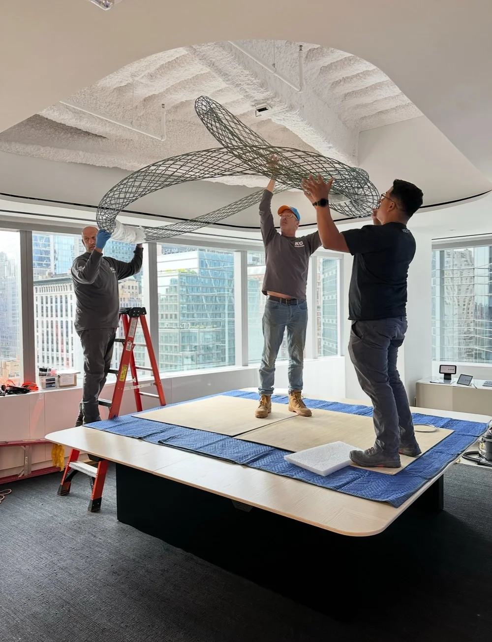 Three art handlers lift a large looped wire sculpture above a work table in an office with city views.