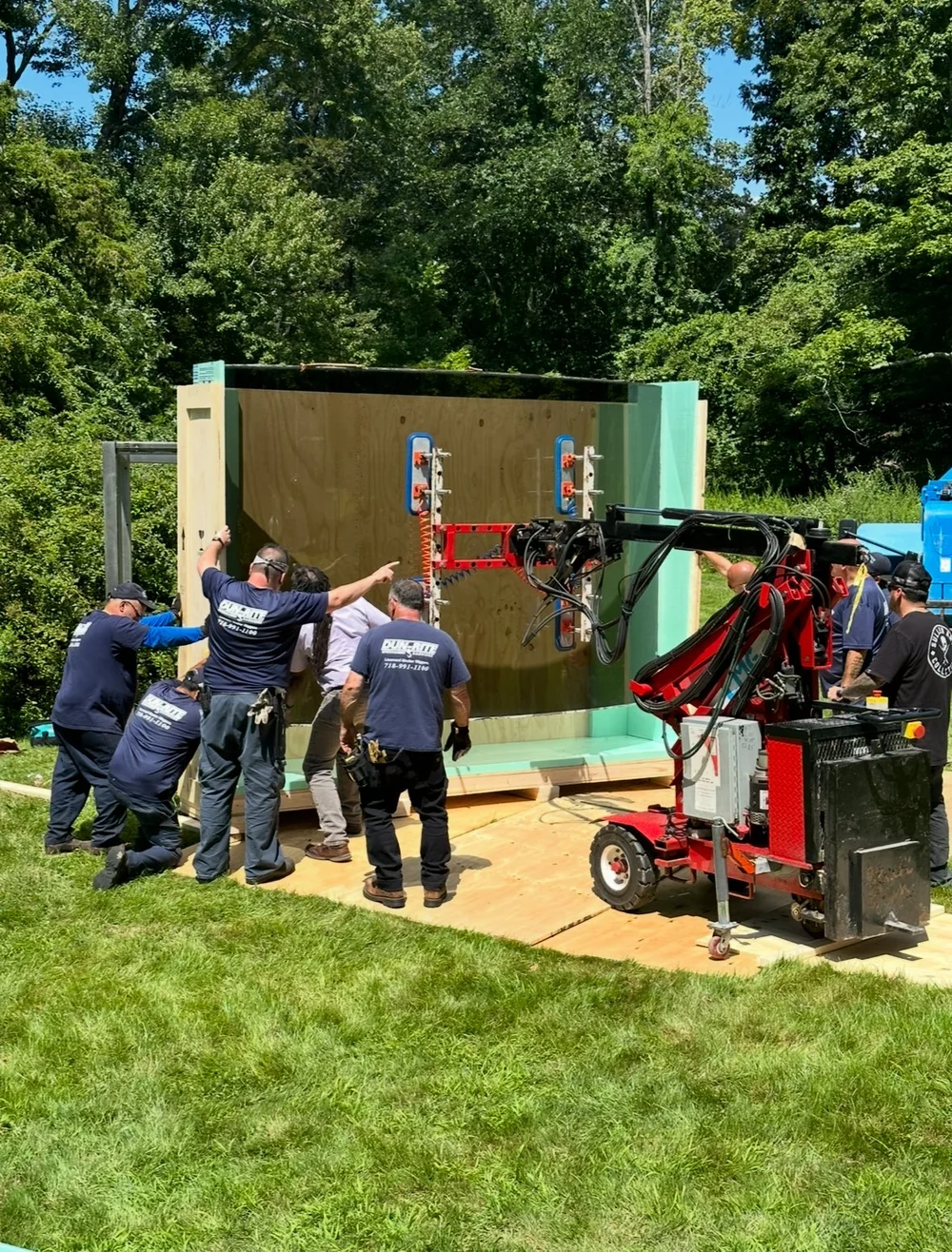 A crew uses a vacuum lifter to move a large crated panel across a lawn, trees behind.