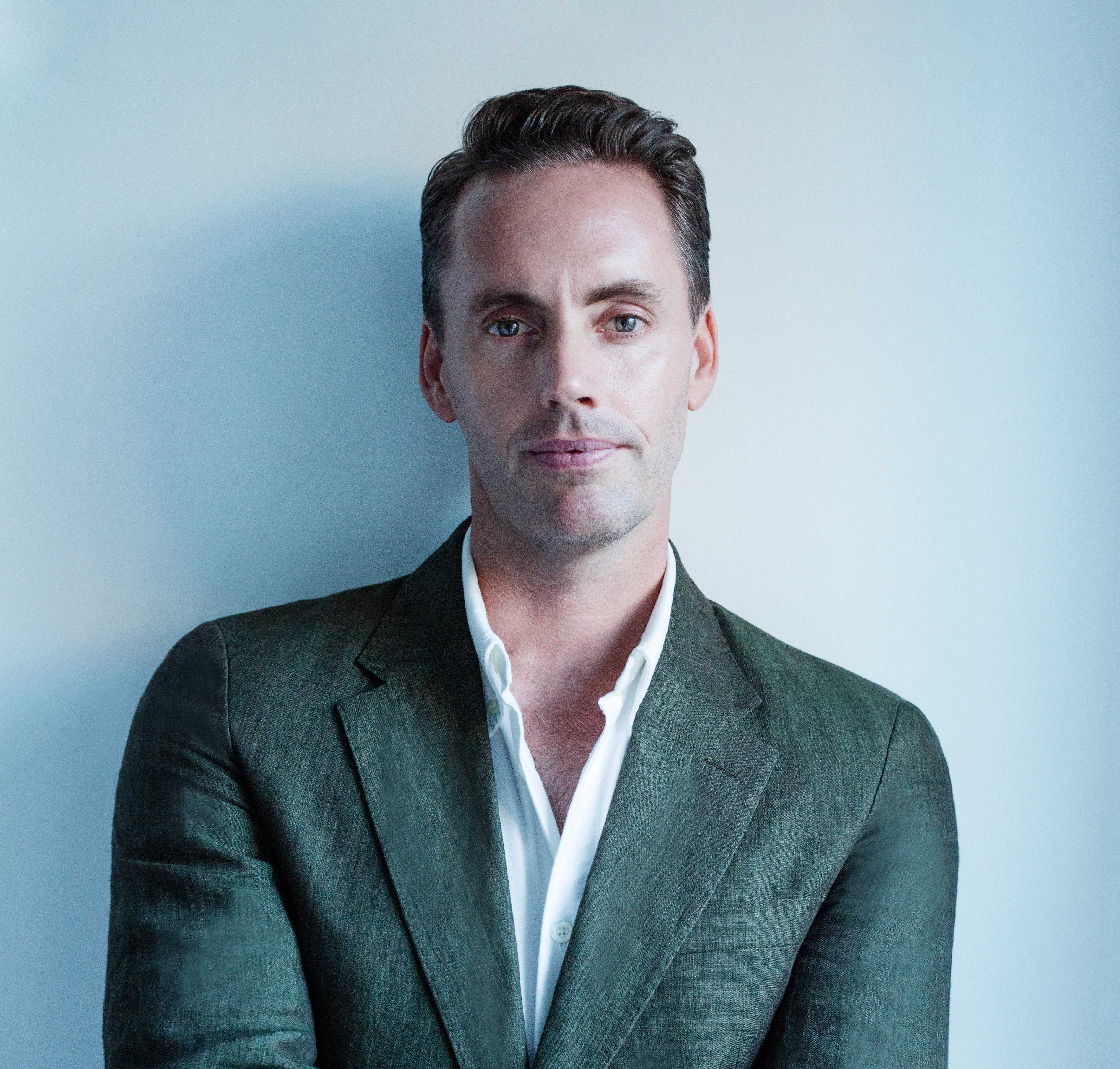 Benjamin Clarke in a green linen jacket and open white shirt against a pale blue wall.